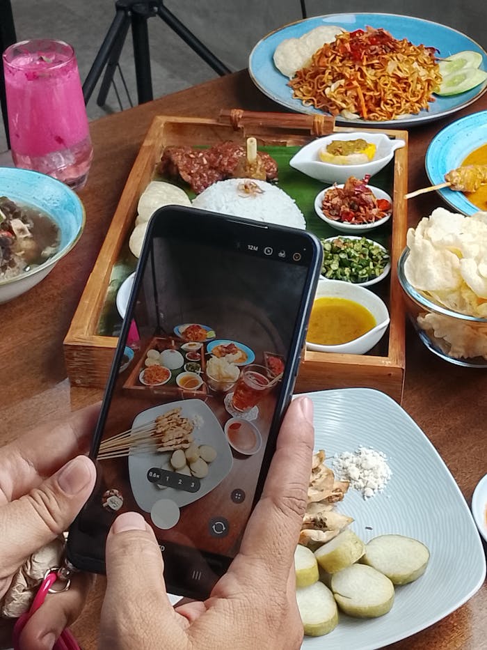 Close-up of a traditional Indonesian meal being photographed on a smartphone, featuring satay and colorful dishes.